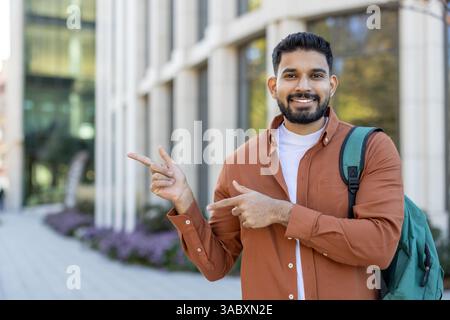 A smiling young man gestures with his hands, pointing towards an area of interest. He is wearing a backpack in front of a modern building. Stock Photo