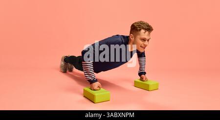 Fit young man doing push-ups with hands on green foam blocks, dressed in navy training outfit, smiling while exercising on pink background. Stock Photo