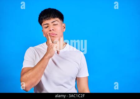 Young man touching his chin, looking down with a thoughtful expression against a vibrant blue background, lost in deep contemplation Stock Photo