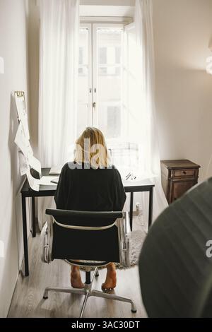 Person sitting at desk in home office, focused on work. Papers are scattered around, suggesting a busy work session. Open window provides natural ligh Stock Photo