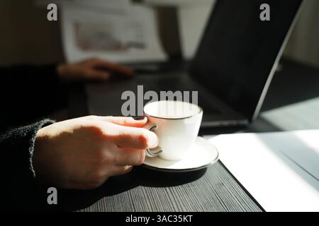 Person holding a teacup while working on a laptop at a desk. The scene suggests a relaxed work session in a quiet, focused setting. Hands are resting Stock Photo