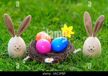 Bavaria, Germany - 2 April 2025: Easter bunnies made of painted wooden eggs with bunny ears stand on a green meadow, surrounded by colorful Easter eggs in an Easter nest and daisies *** Osterhasen aus bemalten Holz Eiern mit Hasenohren stehen auf einer grünen Wiese, umgeben von bunten Ostereiern in einem Osternest und Gänseblümchen Stock Photo