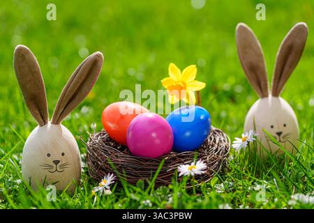 Bavaria, Germany - 2 April 2025: Easter bunnies made of painted wooden eggs with bunny ears stand on a green meadow, surrounded by colorful Easter eggs in an Easter nest and daisies *** Osterhasen aus bemalten Holz Eiern mit Hasenohren stehen auf einer grünen Wiese, umgeben von bunten Ostereiern in einem Osternest und Gänseblümchen Stock Photo