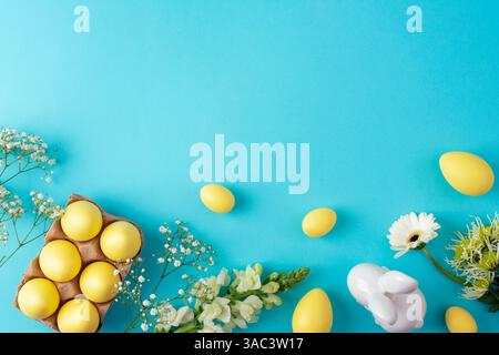 Yellow easter eggs and white spring flowers on blue background with ceramic bunny. Top view, flat lay, copy space. Stock Photo