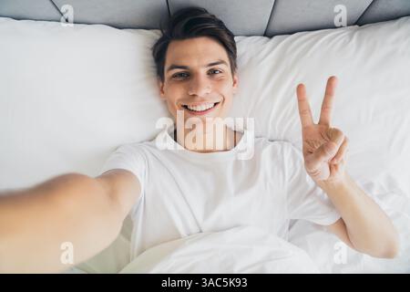 Bright young man in pajama relaxing on bed showing peace sign while smiling Stock Photo