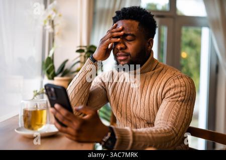 A young black man sits at the bar, using smartphone, messaging online. He is anxious and preoccupied. Stock Photo