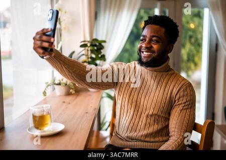 A young black man sits at the bar, casually dressed. He is using smartphone and making selfie. Stock Photo