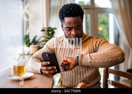 A young black man sits at the bar, cozy dressed. He is using smartphone, messaging online and checking time on his wristwatch. Stock Photo