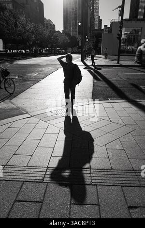 Bathed in morning light, a lone figure waits to cross—while the sun casts a long shadow behind, stretching quietly into the day’s first warmth. Stock Photo