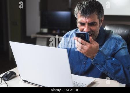 An angry businessman speaking on the phone sitting on a desktop with a laptop. He is talking on the phone while looking at the laptop. Man is in a bad Stock Photo
