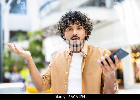 A bewildered young man in a tan shirt holding a phone, looking surprised with his arms open. The background is blurred out. Stock Photo