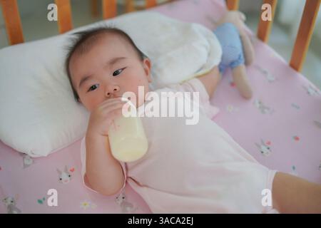 close-up shot of a baby dressed in a pink onesie, comfortably lying on a crib mattress while holding and drinking from a milk bottle. Stock Photo