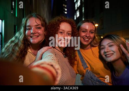 Young female friends taking a selfie looking at camera smiling gathering at night outdoors. Stock Photo