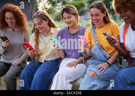 Five cheerful female students are using smartphones while sitting on a bench outdoors in a park. Stock Photo