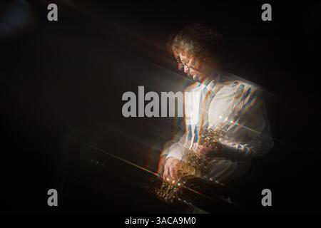 Motion blur shot of adult man in formal attire holding saxophone while standing in spotlight against black backdrop on stage, light dispersion effect Stock Photo