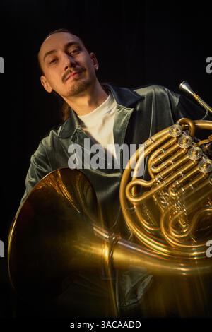 Vertical portrait shot of male jazz player in velvet shirt looking at camera with slight smile while posing with shiny French horn against black backdrop in studio, motion blur effect Stock Photo