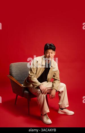 Thoughtful young man sitting in armchair leaning forward slightly with focused look against red backdrop. Concept of contemplation, self-expression Stock Photo