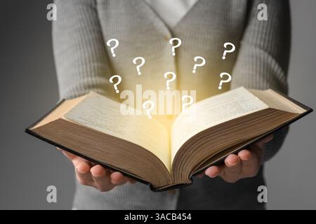Woman holding open book on grey background, closeup. Mysterious light and question marks over pages Stock Photo