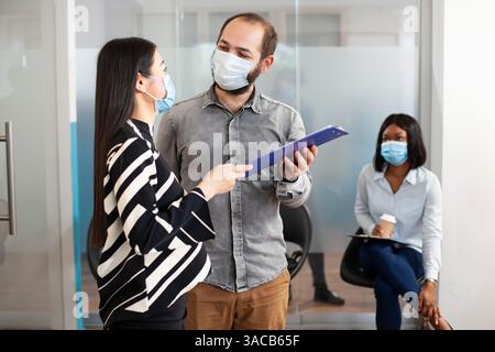 HR male representative invites female candidate to a recruitment meeting, holding her CV during the pandemic. Asian applicant speaking with caucasian man in office lobby, waits for career opportunity. Stock Photo