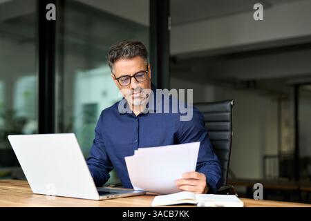 Busy mature professional business man using laptop reading document at work. Stock Photo