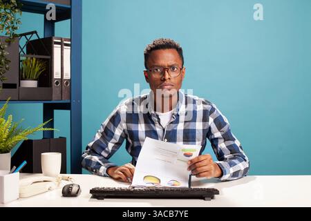 Businessman seated at desk with paperwork, suspicious of the startup project results, having confused expression. POV of male finance analyst checking company accounts on computer, unsure of expenses. Stock Photo