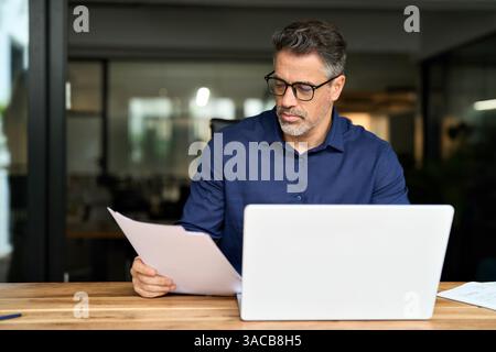 Busy mature professional business man using laptop reading document at work. Stock Photo