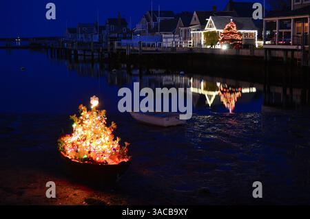 A small illuminated Christmas trees sits in a rowboat while reflecting in the water on the shore of Nantucket island at night Stock Photo