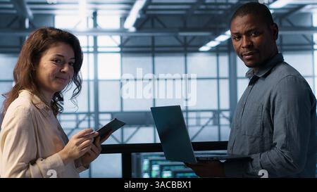 Portrait of happy system administrators in data center using devices, verifying firewall configurations. Jolly teamworking IT experts performing equipment vulnerability scans in server hub, camera A Stock Photo