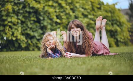 Mother with daughter relaxing on green lawn, sharing peaceful summer moment in backyard Stock Photo