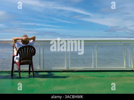 A person relaxes on a chair on the deck of a ferry, gazing out at a clear, tranquil water view under a blue sky with scattered clouds. Stock Photo