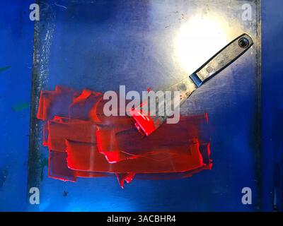 Bright process red ink is spread out on a blue surface with a metal spatula resting beside it, showcasing a creative moment in an artist's studio, rev Stock Photo