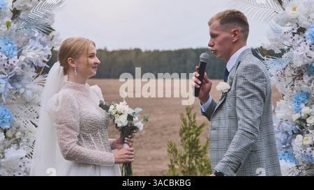 Groom speaking into microphone near bride, standing under decorative floral wedding arch Stock Photo