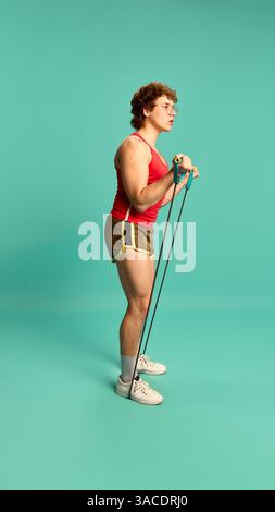 Young man using resistance band for biceps curls, focused and standing firm against mint background studio. Stock Photo