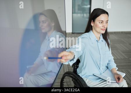 High angle portrait of successful young woman in wheelchair leading business meeting with team and pointing at whiteboard, copy space Stock Photo