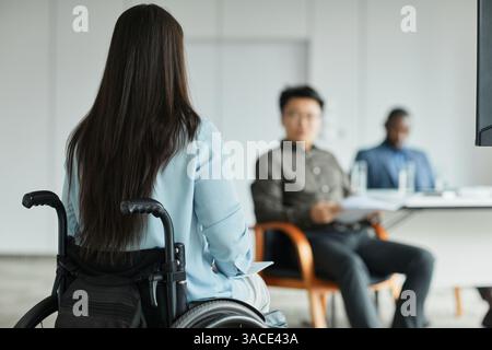 Back view portrait of young woman in wheelchair talking to diverse business team during group discussion in office, copy space Stock Photo