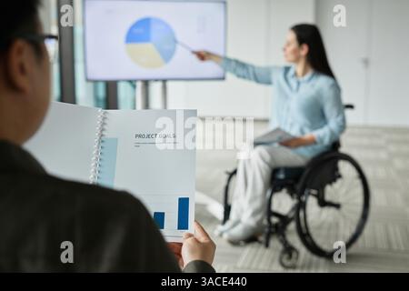 Blurred shot of businesswoman in wheelchair leading team meeting and pointing at digital whiteboard with focus on Project goals planner in foreground, Stock Photo
