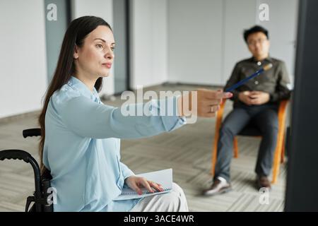 Portrait of successful businesswoman in wheelchair pointing at whiteboard while giving report or leading team meeting in office Stock Photo
