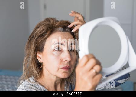 Gray haired shocked caucasian middle aged mature woman looking at grey hair head in mirror reflection at home. Concept of age-related changes, natural Stock Photo