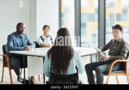 Back view portrait of young woman in wheelchair collaborating with to diverse business team in office, copy space Stock Photo