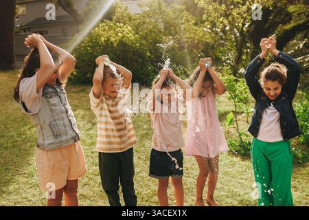 A group of children have fun splashing water on each other in the yard. The scene captures joy, friendship, and playfulness, in the sunlight Stock Photo