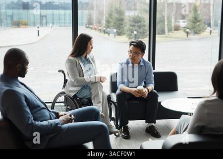 Diverse group of business people chatting in office lobby with focus on young woman in wheelchair Stock Photo