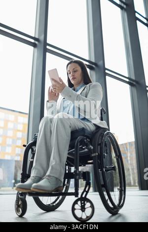 Vertical low angle portrait of young businesswoman in wheelchair using smartphone in office lobby Stock Photo