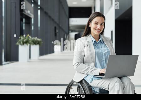 Portrait of successful young businesswoman in wheelchair using laptop and smiling at camera in office lobby, copy space Stock Photo