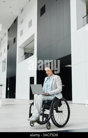 Vertical wide angle portrait of successful businesswoman in wheelchair using laptop in minimal office lobby, copy space Stock Photo