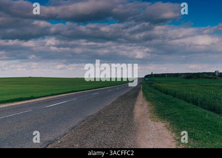 Empty Country Road Through Green Fields Under Cloudy Sky Stock Photo