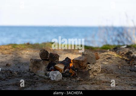 A Serene Lakeside Campsite Featuring a Cozy Campfire Surrounded by Rocks Against a Backdrop of Calm Water and Lush Greenery Beneath a Clear Blue Sky Stock Photo