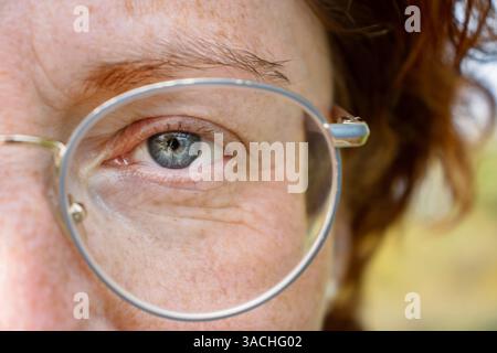 Detailed closeup of the left  blue gray eye of a serious middle aged red haired woman wearing glasses in metal frame. Half of the face  Macro shot. Fi Stock Photo