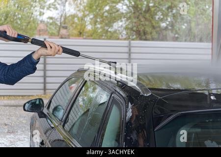 The image shows a person using a pressure washer to clean the roof of a black car at a car wash facility. The metal fence and trees in the background Stock Photo