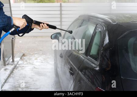 The image shows a person using a high-pressure water hose to clean a black car covered in soap and water. Stock Photo