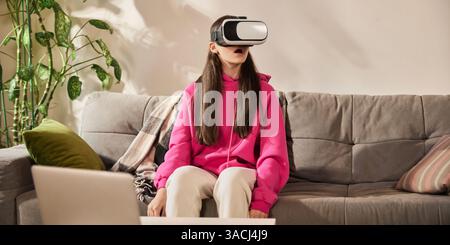 Shocked and excited teen girl in pink hoodie amazed while exploring simulation through VR headset during interactive lesson at home. Stock Photo